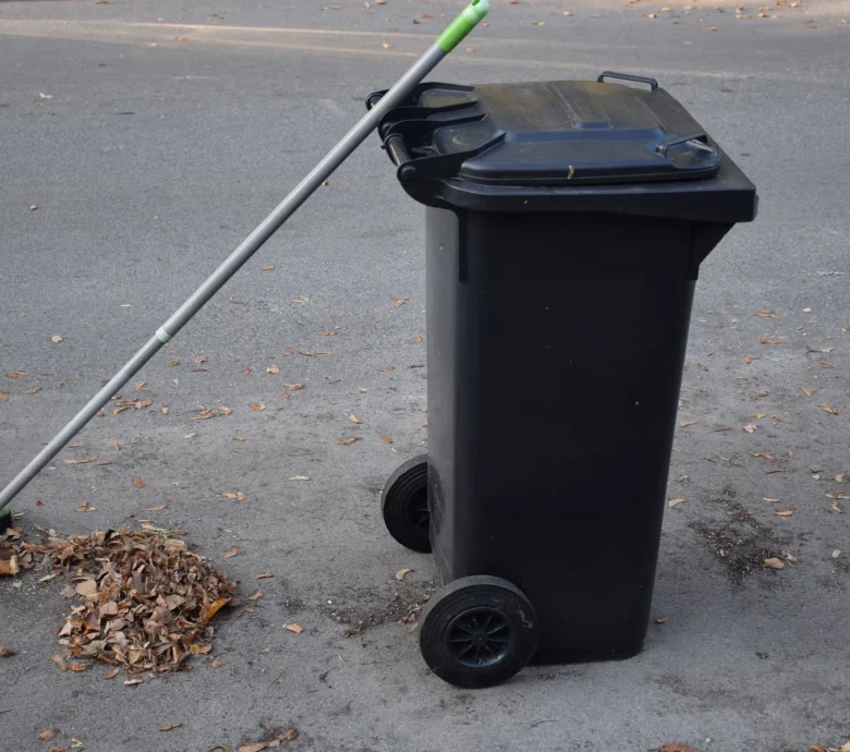 Černý kontejner na odpadky s koštětem vedle hromady listí.