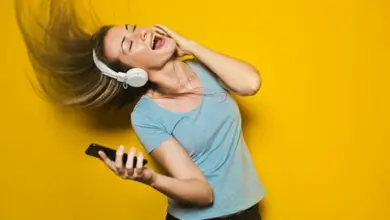 Žena s bezdrátovými sluchátky drží telefon na žlutém pozadí.