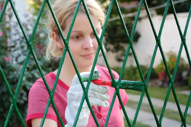 Žena v rukavicích natírá zelenou barvou drátěný plot štětcem.