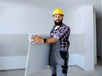 Muž v žluté helmetu drží sádrokartonovou desku.