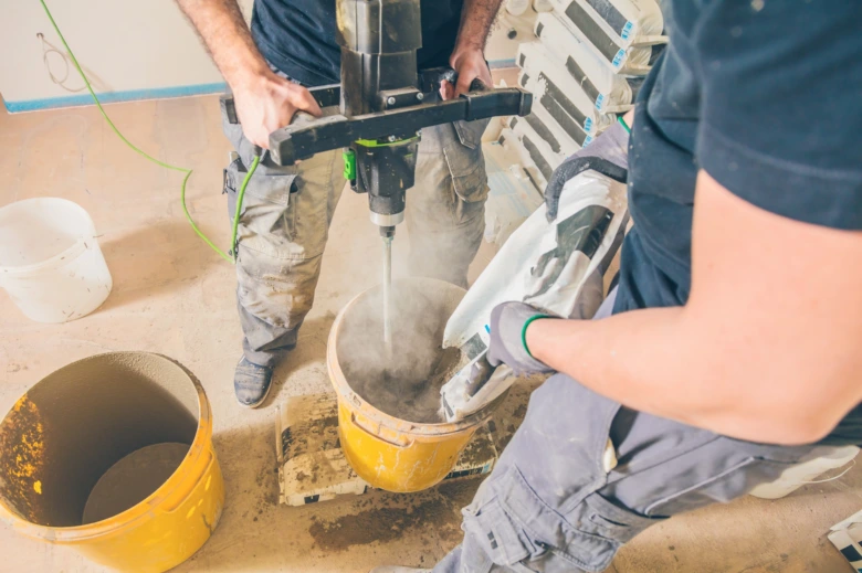 Dva dělníci míchají beton v kbelíku pomocí elektrického nástroje.