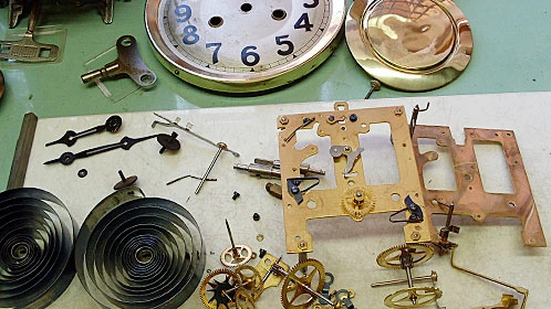 Rozložené díly mechanických nástěnných kyvadlových hodin na pracovní ploše.