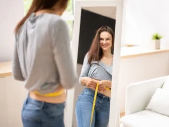 Žena měří pas žlutou páskou před zrcadlem v džínách.