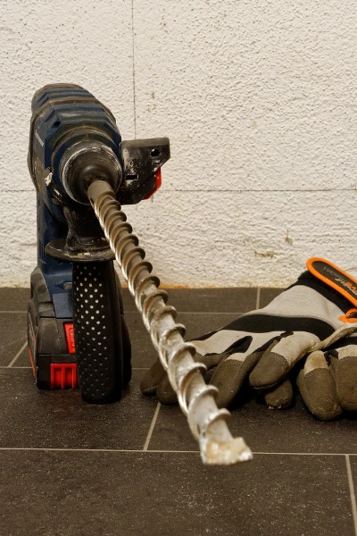 Modrý vrták s dlouhou bity leží vedle pracovních rukavic na dlažbě.