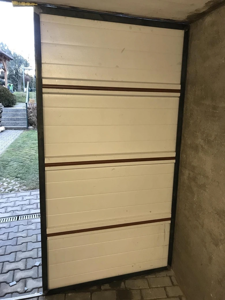 Část bílých garážových vrat s hnědými pruhy polootevřená venku.