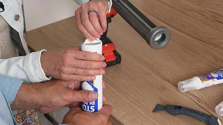 Ruce aplikují bílý silikonový tmel z pistole na dřevěnou plochu.