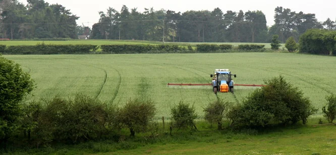 Traktor postřikuje pole v krajině s stromy.