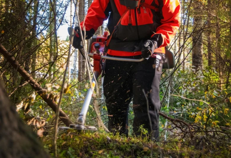 Osoba v ochranném obleku řezá keře krovinorezem ve lesu.