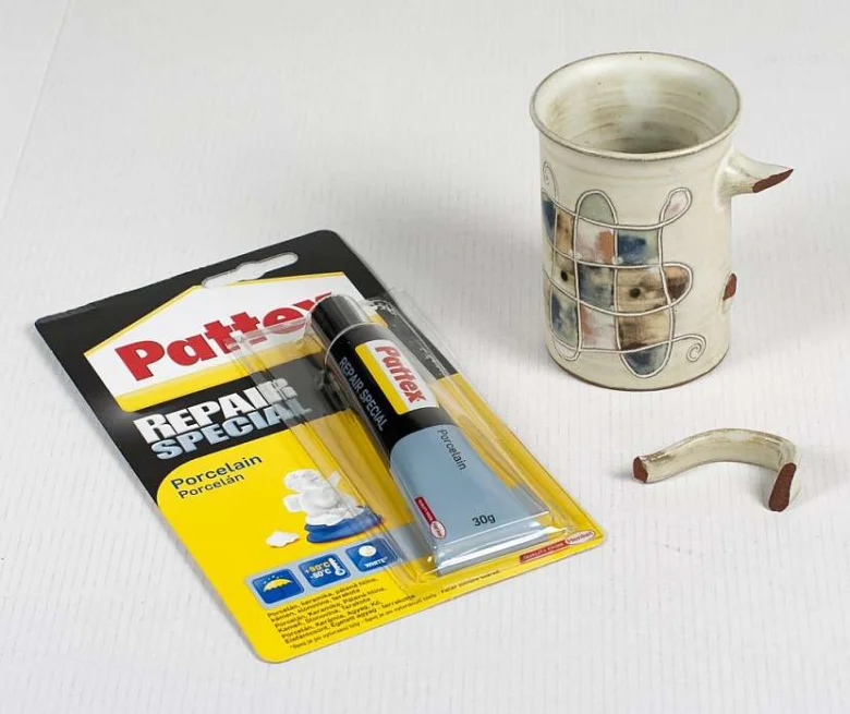 Sada na opravu porcelánu vedle rozbitého hrnku s odlomenou rukojetí.