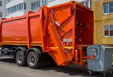 Oranžový nákladní vůz sběru odpadu zvedá kontejner u domu.