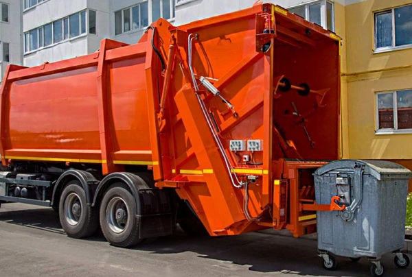 Oranžový nákladní vůz sběru odpadu zvedá kontejner u domu.