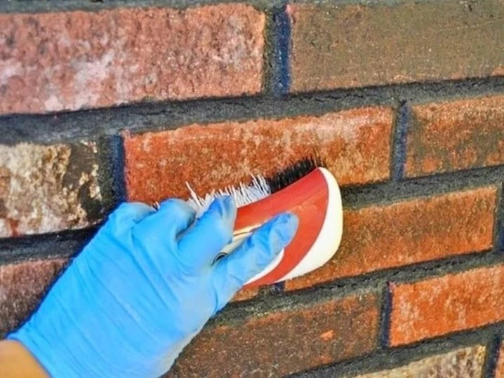 Ruka v modré rukavici čistí cihlovou zeď kartáčem.
