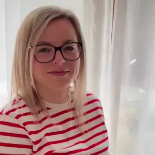Žena v brýlích s pruhovaným tričkem před záclonou.