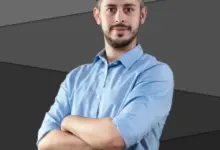 Muž v modré košili s kříženýma rukama na tmavém pozadí.