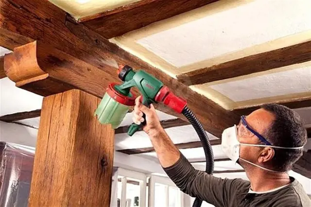 Muž stříká dřevěný trám na strop pomocí stříkací pistole.