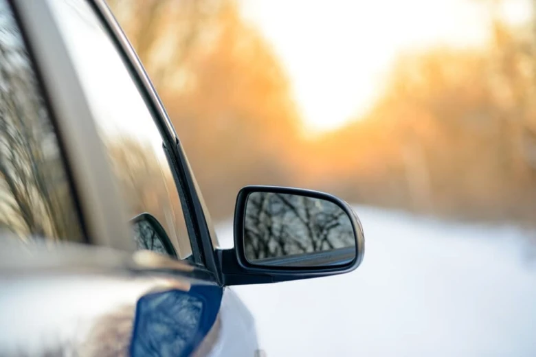 Bílý automobil v zimě s odrazem stromů ve zrcátku.