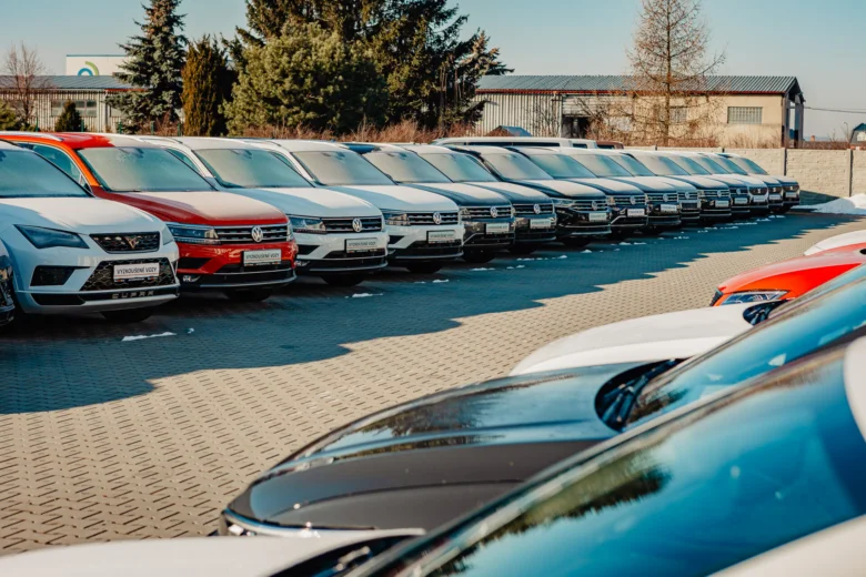 Řada nových osobních automobilů zaparkovaných na asfaltovém parkovišti.
