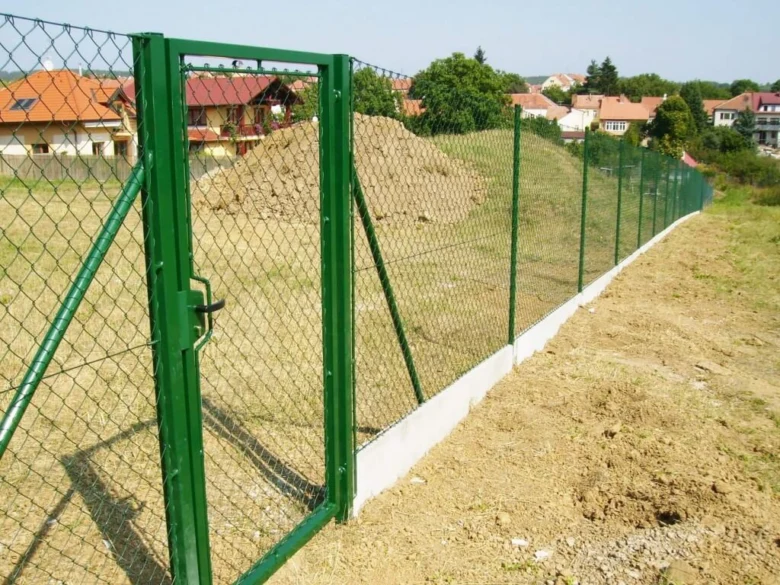 Zelený plot s bránou na pozadí domů.
