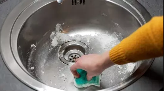 Ruka čistí nerezový dřez houbou od špíny.