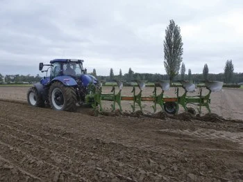 Modrý traktor s pluhem na poli při orání půdy.
