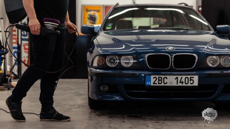 Muž brousí modrý automobil v dílně pomocí elektrického polírovacího stroje.