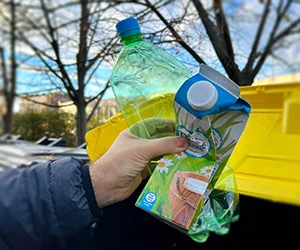 Ruka drží zelenou PET lahvičku vedle kartonového balení u žlutého kontejneru na recyklaci.