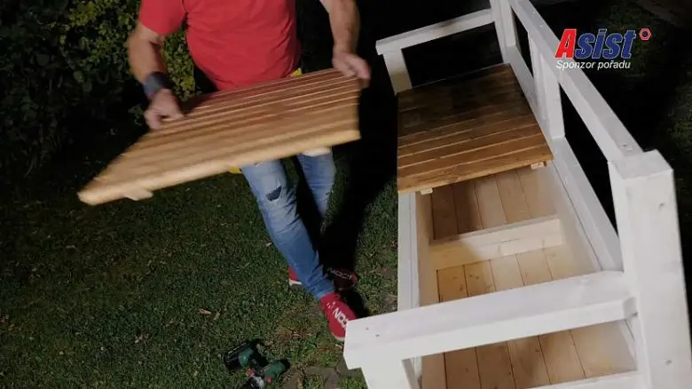 Muž sestavuje dřevěnou lavičku s bílým rámem venku.