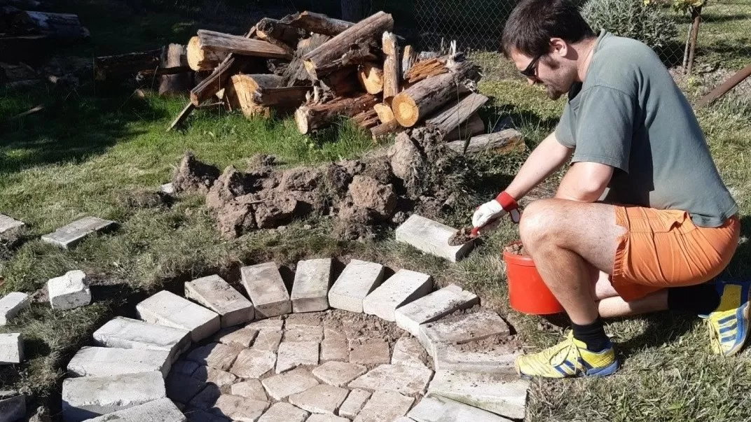 Muž staví kruhový krb z cihel na trávníku.