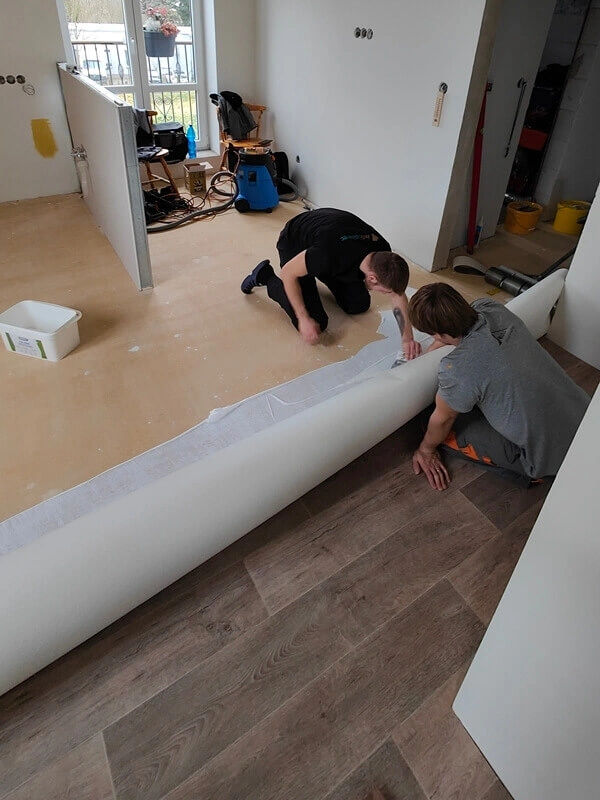 Muži narovnávají podklad pro linoleum bez vrásek.